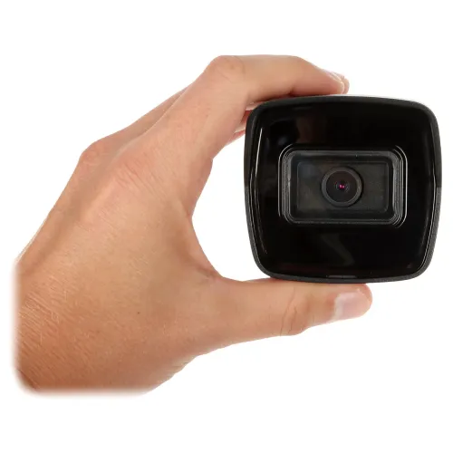 Na zdjęciu widać dłoń trzymającą niewielką kamerę samochodową, skierowaną obiektywem w stronę widza. Kamera ma prostokątny kształt i czarną obudowę.