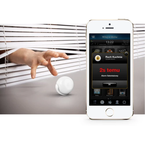 Na zdjęciu znajduje się biały, kulisty czujnik ruchu umieszczony na płaskiej powierzchni, obok którego widoczny jest smartfon z aplikacją pokazującą powiadomienie o wykrytym ruchu. Czujnik jest prezentowany w kontekście inteligentnego systemu domowego, co sugeruje jego funkcje związane z bezpieczeńs