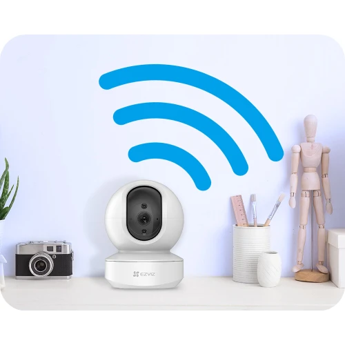 Kamera IP Ezviz TY1 w kolorze białym, umieszczona na biurku, z widocznym obiektywem i logo producenta. W tle znajduje się niebieska ikona sygnału Wi-Fi oraz drewniana figurka i przybory biurowe.