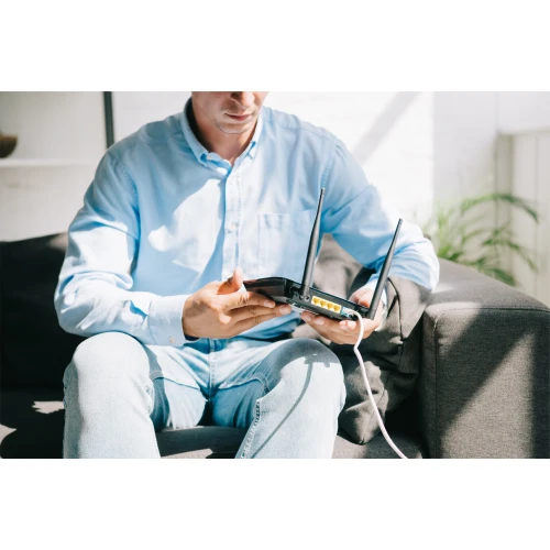 Mężczyzna w niebieskiej koszuli siedzi na kanapie, trzymając router z widocznymi antenami i portami. W tle widać jasne wnętrze z rośliną.